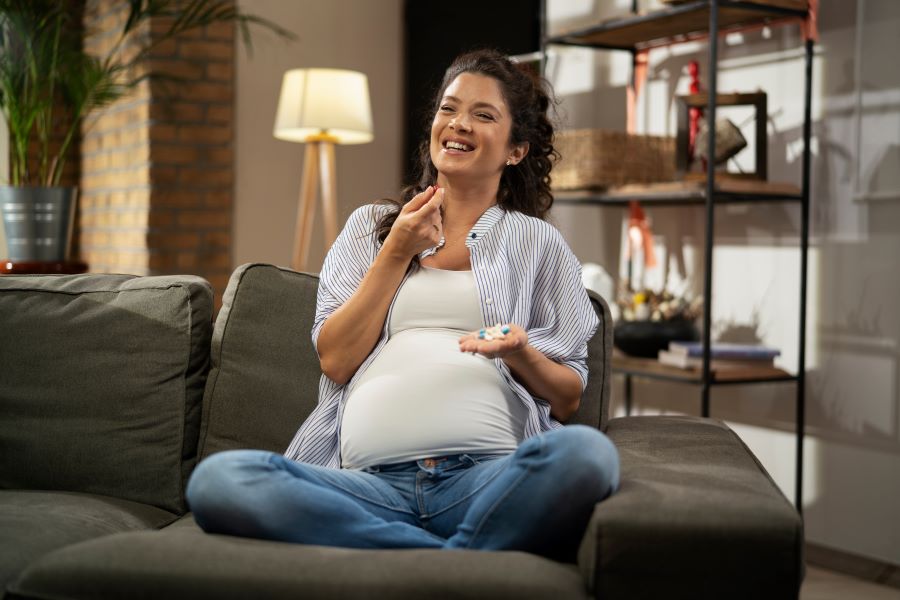 A pregnant women smiles while sitting on the couch and preparing to take a supplement. A pregnant women smiles while sitting on the couch and preparing to take a supplement.