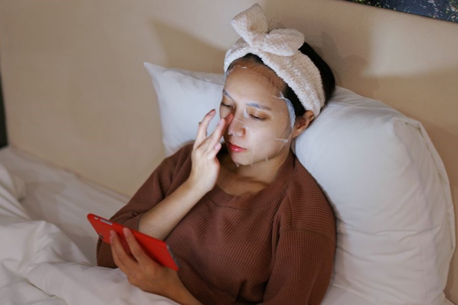 Woman laying in bed wearing sheet mask on her face and watching her phone Woman laying in bed wearing sheet mask on her face and watching her phone
