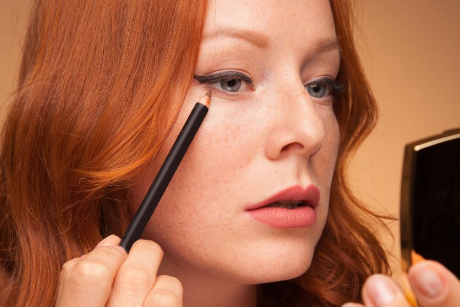 Woman applying eyeliner pencil while looking in compact mirror
