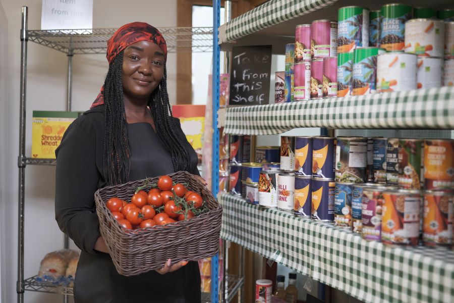 Black woman holds a basket of tomatos in next to shelves of canned food