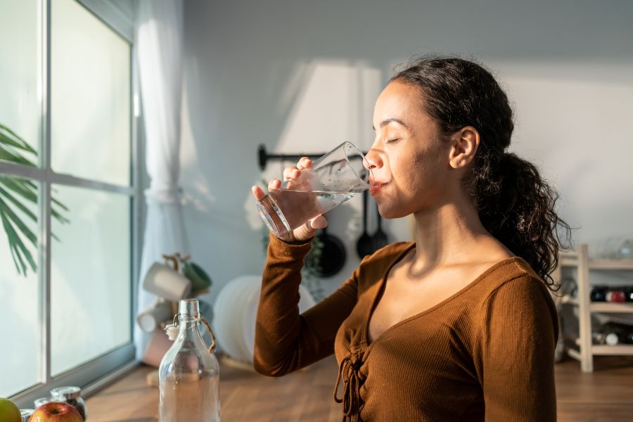 A woman drinking water on a sunny day in front of the kitchen sink.