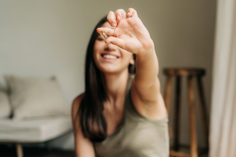 A woman happily holds up an omega-3 supplement while sitting on a couch.