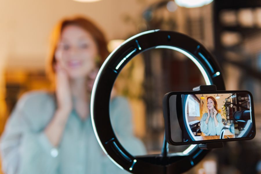 A woman putting a roller on her face as seen through her phone in front of a ring light.