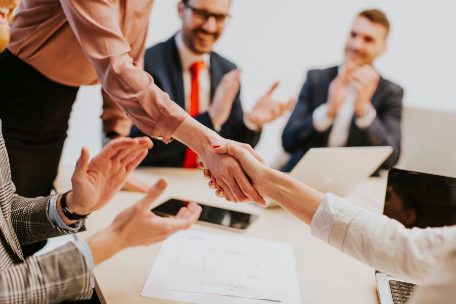 A group of people in a bright business office applaud as two women shake hands on a business deal.