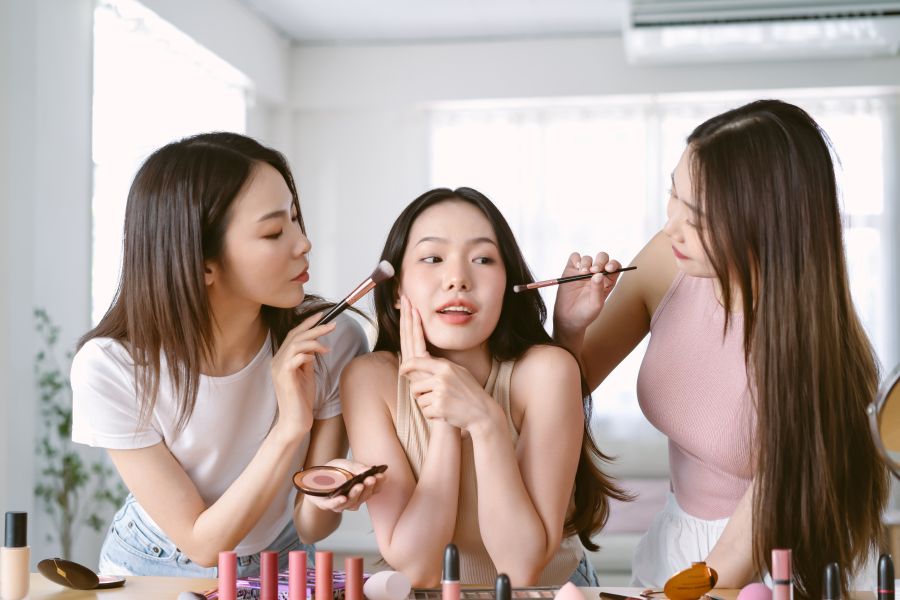 Two girls doing the makeup of a girl in the middle of them.