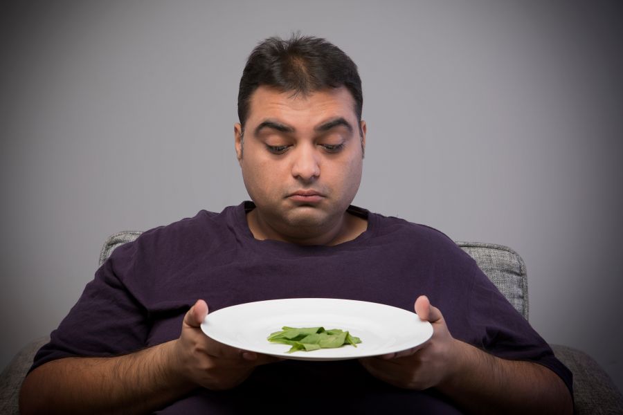 An overweight man stares sadly at a couple of peices of lettuce on the plate he is holding at the dinner table. An overweight man stares sadly at a couple of peices of lettuce on the plate he is holding at the dinner table.