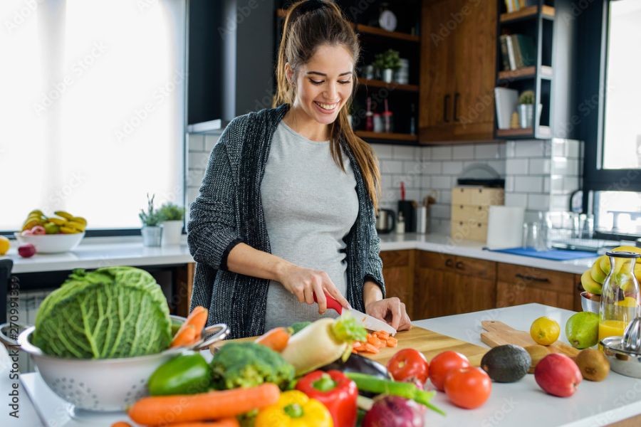 A woman chops up vegetables on a cutting board that rests on a countertop that is covered with vegetables and fruits.
