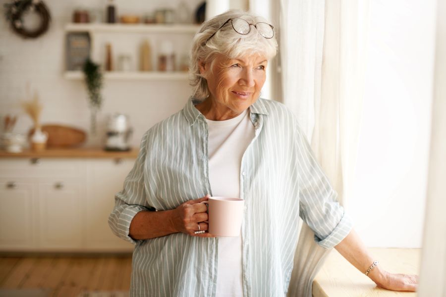 Portrait of stylish gray haired woman enjoying a drink, holding mug, looking outside through window