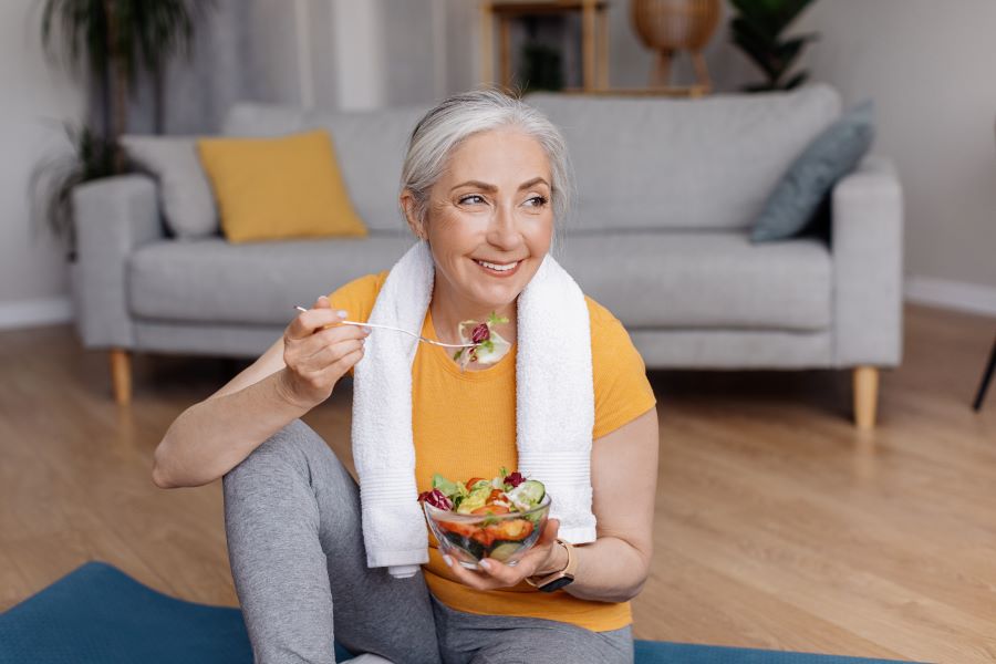 Happy senior woman eating fresh vegetable salad, sitting on yoga mat after home workout