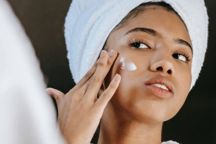 A woman putting lotion on her cheek while her hair is in a towel.