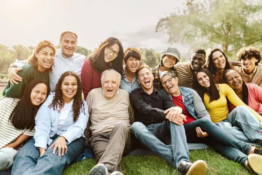Many people of different ages and demographics all sitting happily in the grass at a park.
