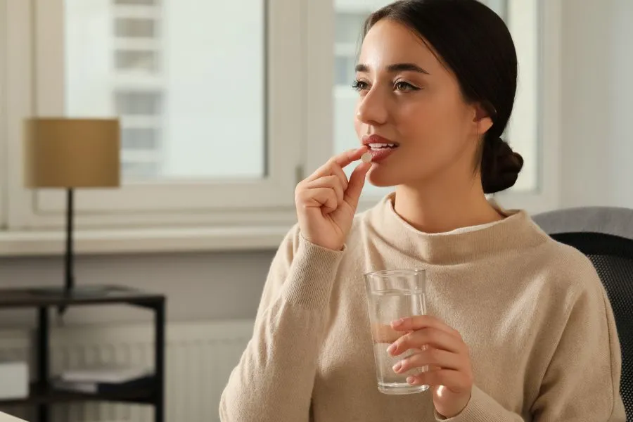 A woman taking a supplement while wearing a beige sweater and sitting on a couch.