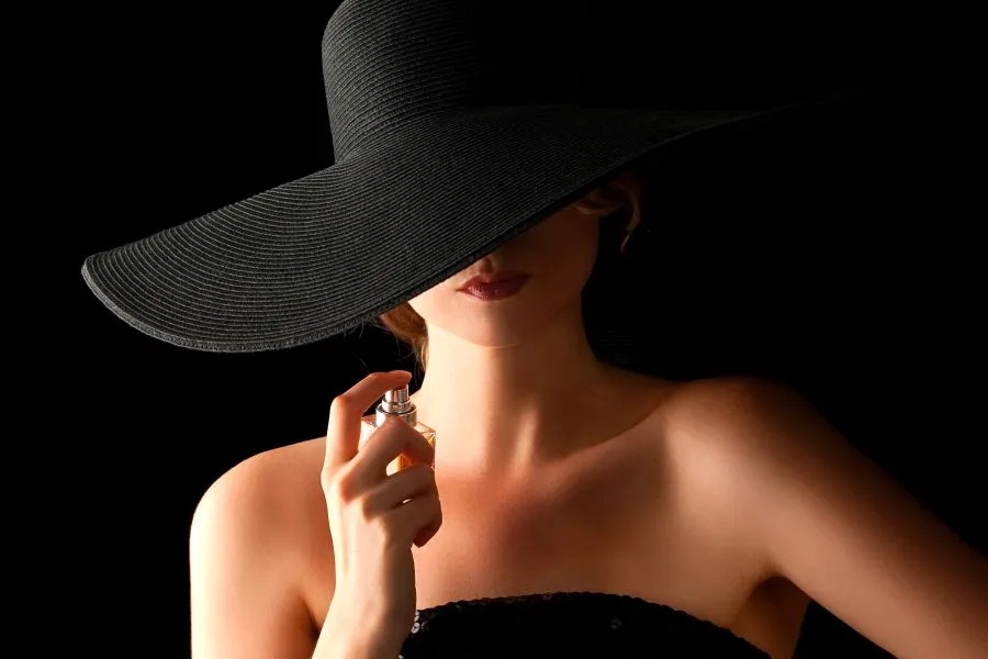 A woman wearing a wide hat covering her face and holding a bottle of perfume.
