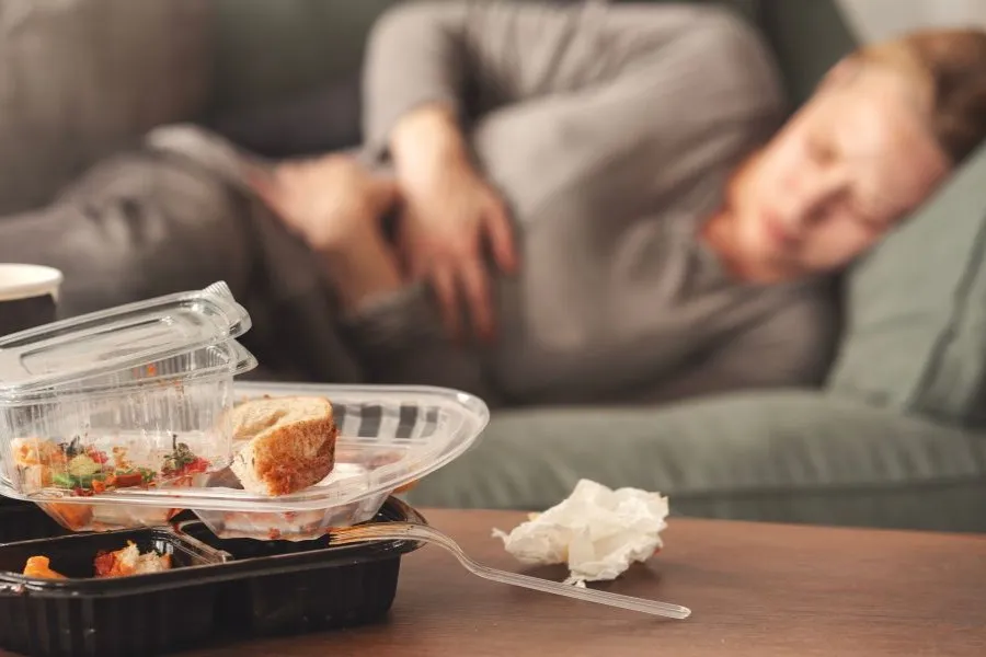 A woman clutches her stomach while lying on the couch with empty cartons of take out food on the table in front of her.