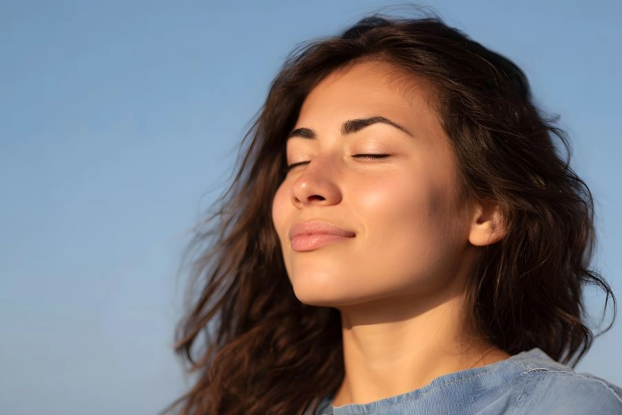 Young woman in golden hour sunlight eyes closed taking a deep peaceful breath under a clear blue sky