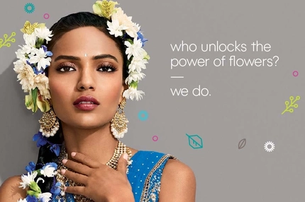 Woman wearing blue traditional attire adorned with white and blue flowers in her hair, statement gold jewelry, and soft glam makeup, with floral-themed branding text beside her.