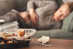A woman clutches her stomach while lying on the couch with empty cartons of take out food on the table in front of her.