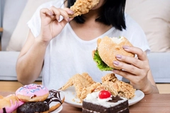 woman in white shirt biting into fried chicken with burger and donuts on table