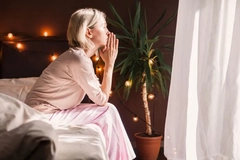 A woman sitting on the bed with her hands on her chin looking out a window.