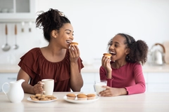 A mother and daughter eating cookies and smiling at a table in the kitchen.