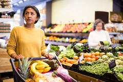 Pensive latina buying groceries at store, walking with shopping cart among shelves and choosing products