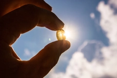 Hand holding soft gel fish oil or vitamin d capsule against blue sky and sun.